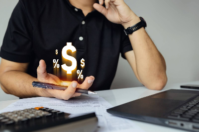 persona analizando documentos financieros, con una calculadora, una laptop y un bolígrafo sobre el escritorio. De su mano derecha surge un efecto visual de luz con símbolos de dólar y porcentajes