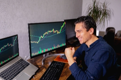 Esta imagen muestra a un hombre celebrando con entusiasmo frente a sus pantallas de trading, las cuales muestran una clara tendencia alcista.
