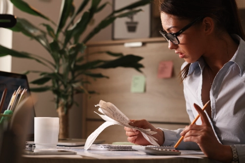 Una mujer con gafas revisando concentrada un fajo de facturas largas mientras utiliza una calculadora