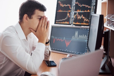 un joven analista financiero en un momento de intensa reflexión o estrés. Está sentado frente a un escritorio con múltiples monitores que muestran gráficos de velas japonesas y tendencias de mercado complejas