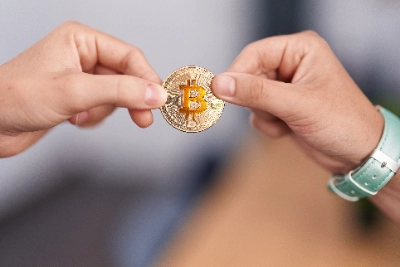 Dos personas sostienen e intercambian una moneda física dorada de Bitcoin, simbolizando una transacción o transferencia de criptomonedas.
