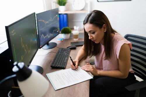 Una mujer de negocios firma un contrato físico en su escritorio, junto a dos monitores que muestran gráficos financieros.