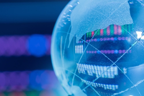 Primer plano de un globo terráqueo de cristal translúcido que refleja en su superficie gráficos financieros de velas y datos numéricos borrosos en tonos azules, verdes y rojos.