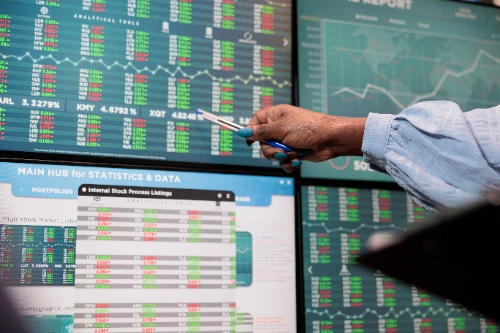 Primer plano de una mano señalando con un bolígrafo una pantalla repleta de datos, tablas numéricas y gráficos estadísticos del mercado financiero.