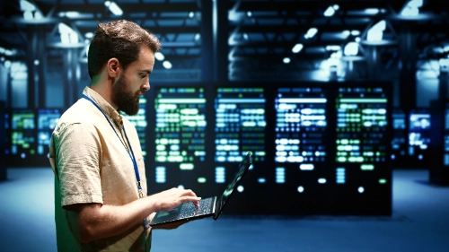 se muestra de perfil a un hombre con barba que utiliza una computadora portátil mientras trabaja de pie en una sala de servidores iluminada con luces azules y verdes.