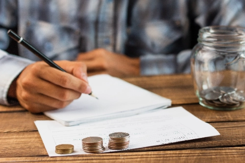 Primer plano de una mano escribiendo con lápiz junto a varias pilas de monedas y un frasco de vidrio para ahorros en una mesa de madera.