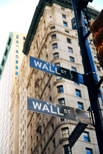 se muestran dos carteles callejeros negros que indican "WALL ST" en un poste en primer plano, con la fachada de un edificio clásico de Nueva York de fondo en un plano contrapicado.