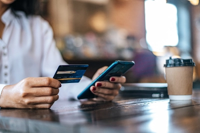 una mujer sostiene una tarjeta de crédito en una mano y un teléfono inteligente en la otra, sentada a una mesa de madera en una cafetería junto a un vaso de café para llevar.
