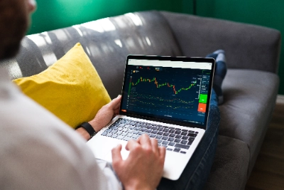 una perspectiva sobre el hombro a un hombre recostado en un sofá gris con un cojín amarillo, utilizando una laptop que muestra gráficos del mercado financiero.