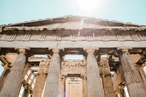 La fachada de un antiguo templo de piedra con grandes columnas se alza bajo un cielo claro. El sol brilla con fuerza en la parte superior, creando destellos de luz.
