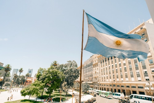 La bandera de Argentina ondea en un mástil en primer plano. Al fondo se observa una plaza con árboles, edificios y calles de la ciudad.