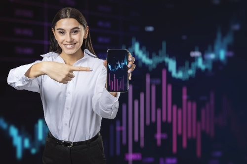 Una mujer sonriente señala la pantalla de su teléfono móvil, la cual muestra gráficos de velas financieras sobre un fondo digital con barras y curvas neón difuminadas.