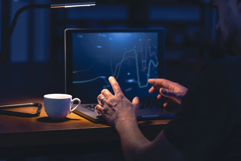 Un hombre analiza gráficos de líneas en la pantalla de su portátil de noche, con una taza de café al lado sobre el escritorio iluminado por una lámpara.