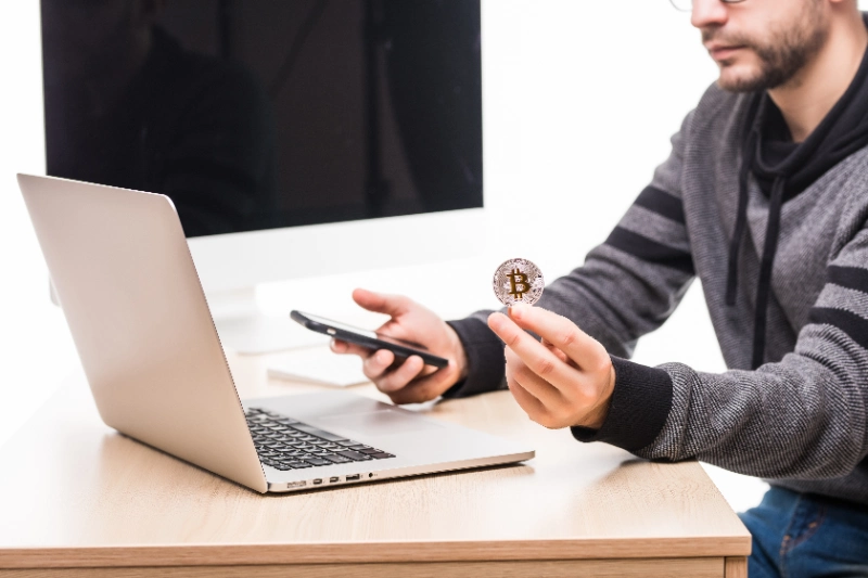 Un profesional sentado frente a un ordenador portátil sostiene una moneda física de bitcoin y un teléfono móvil sobre un fondo neutro.