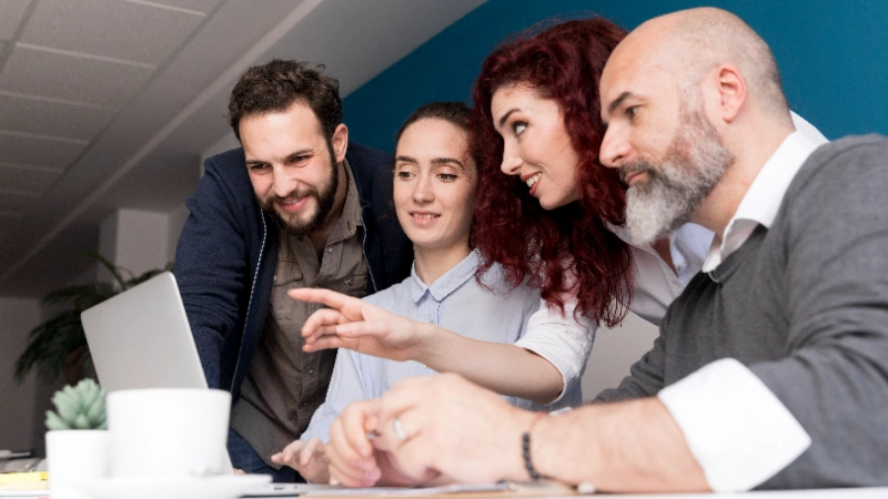 Equipo diverso de profesionales reunido alrededor de una laptop en la oficina mientras observan y comentan el contenido de la pantalla.