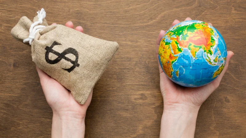 Manos sosteniendo un saco de dinero de arpillera con el signo de dólar en un lado y un globo terráqueo en el otro sobre una superficie de madera.