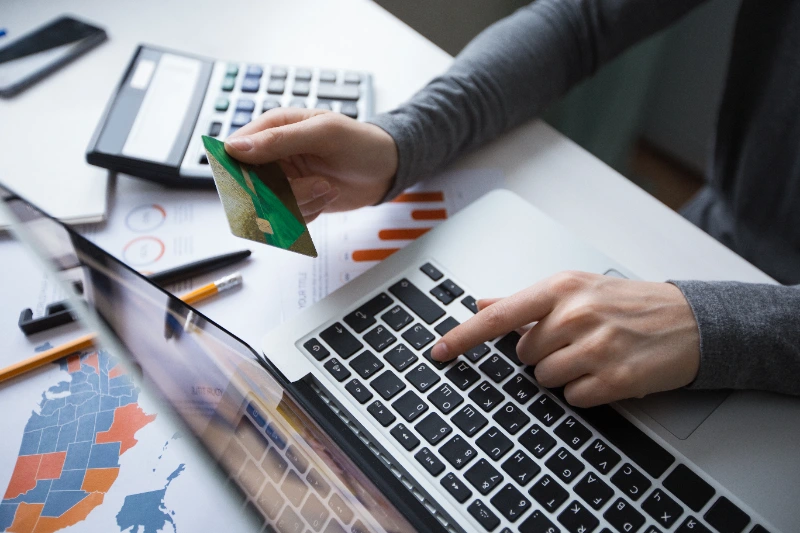 Vista de un gestor realizando un pago en línea con tarjeta de crédito en una computadora portátil rodeada de informes financieros y calculadora.