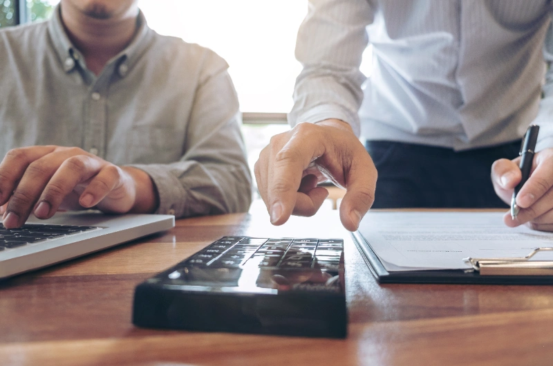 Dos profesionales colaborando en una mesa de trabajo, uno de ellos utilizando una calculadora mientras el otro opera una computadora portátil y revisa documentos corporativos.